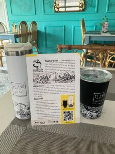 A reusable tumbler and mug, alongside a paper explaining the project, sit on a counter.